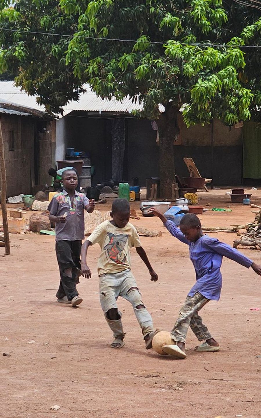 Enfants jouant au ballon sur une place de terre battue, devant des habitations et un manguier